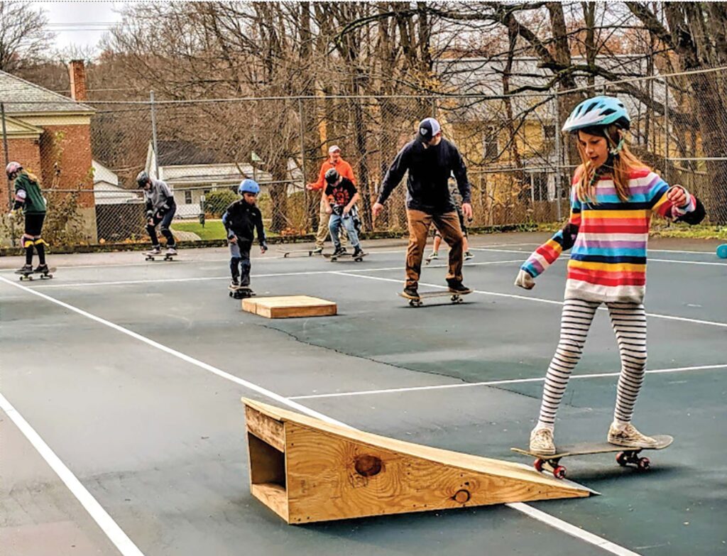 A mixed-age group practices skateboarding at an interim skatepark, on a tennis court with wooden ramps.