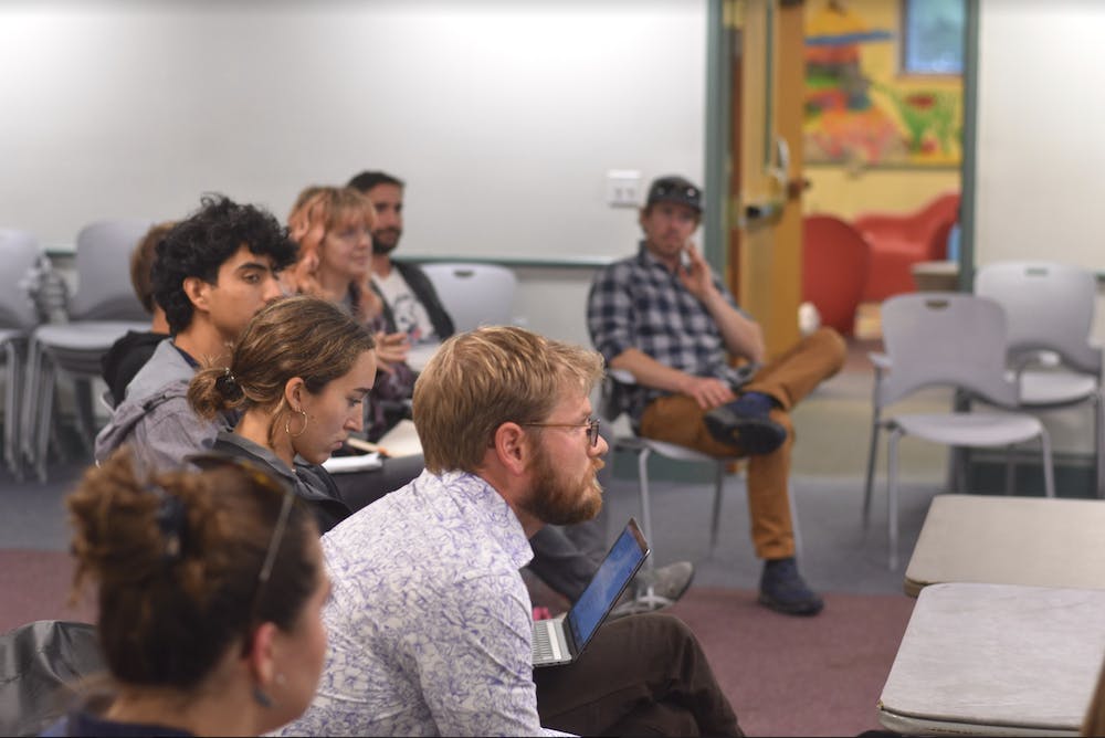 A group of people, ranging from college students to middle age parents, sits together in a meeting room listening intently to a speaker who is not visible.