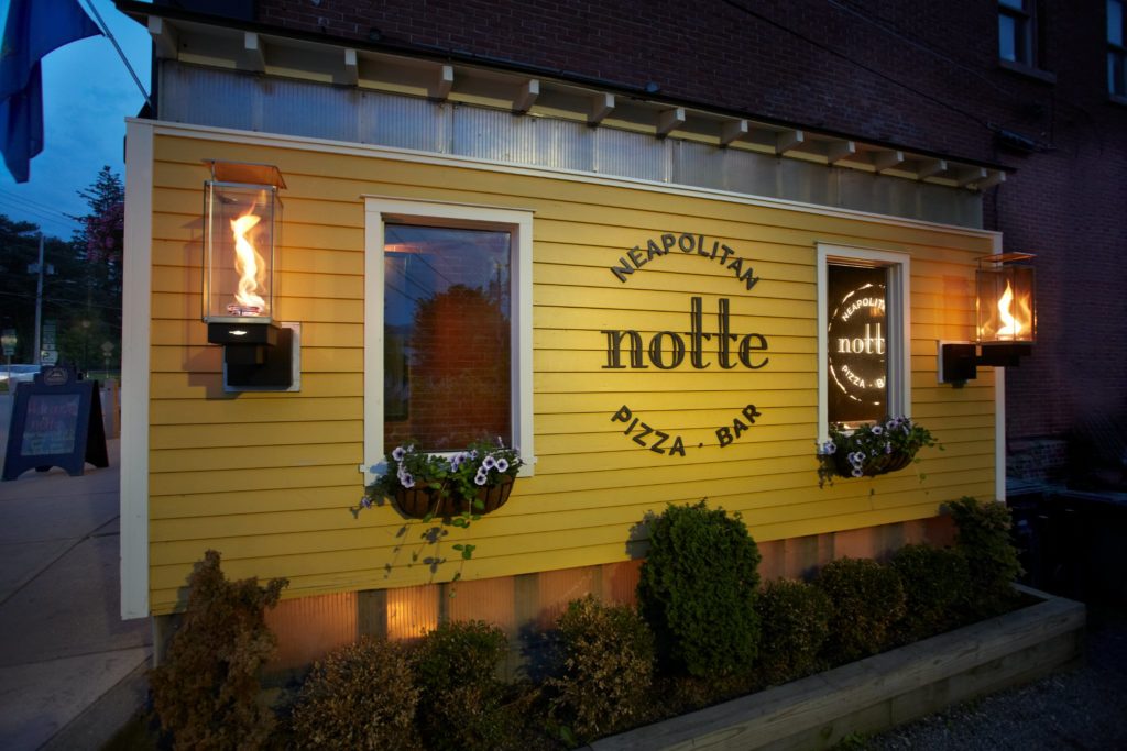 Nighttime exterior photo of Notte, a yellow friendly-looking building with sign saying "Notte, neopolitan pizza and bar". The sign is flanked by two windows with flower boxes and two gas lanterns.