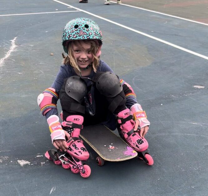 A young girl is wearing a helmet, pads, and roller blades and is sitting on a skateboard. She is smiling and having fun rolling around on ALL her wheels!